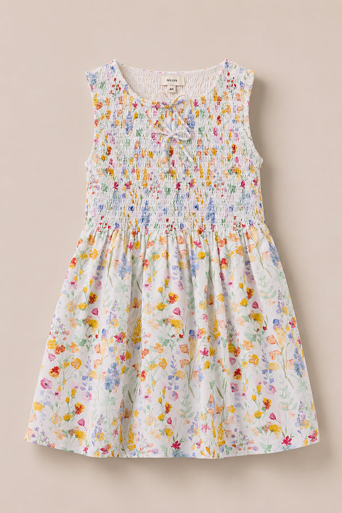 The Mini Ana Dress in Floral Fields by Ivy City Co is a sleeveless, floral print dress for girls with a smocked bodice and a bow at the neckline, shown on a light beige background.