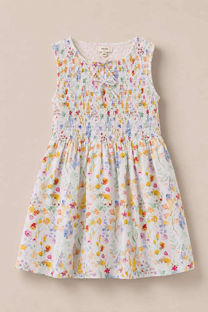 The Mini Ana Dress in Floral Fields by Ivy City Co is a sleeveless children’s dress featuring a smocked, ruched bodice with a delicate neck tie. Its skirt displays a vibrant floral pattern on a light background, making it the perfect floral dress for kids.