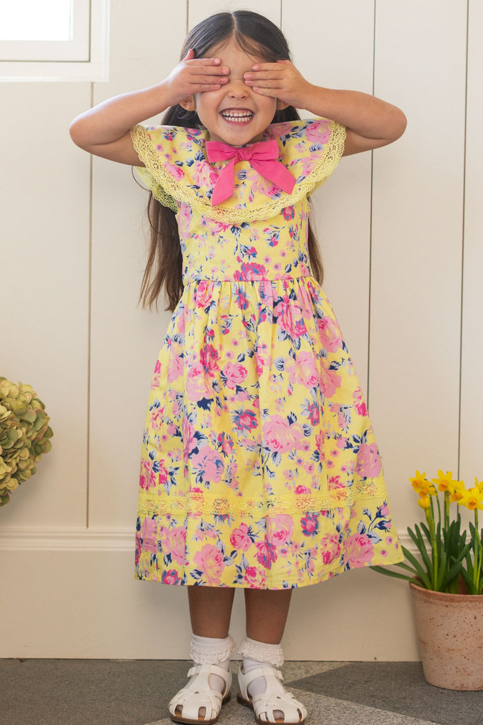 A young girl in Ivy City Co’s Mini Aster Dress in As You Dream Floral, with a pink bow, smiles and covers her eyes indoors by a white paneled wall. Potted flowers on the floor beside her echo the cheerful vibe of her floral dress.