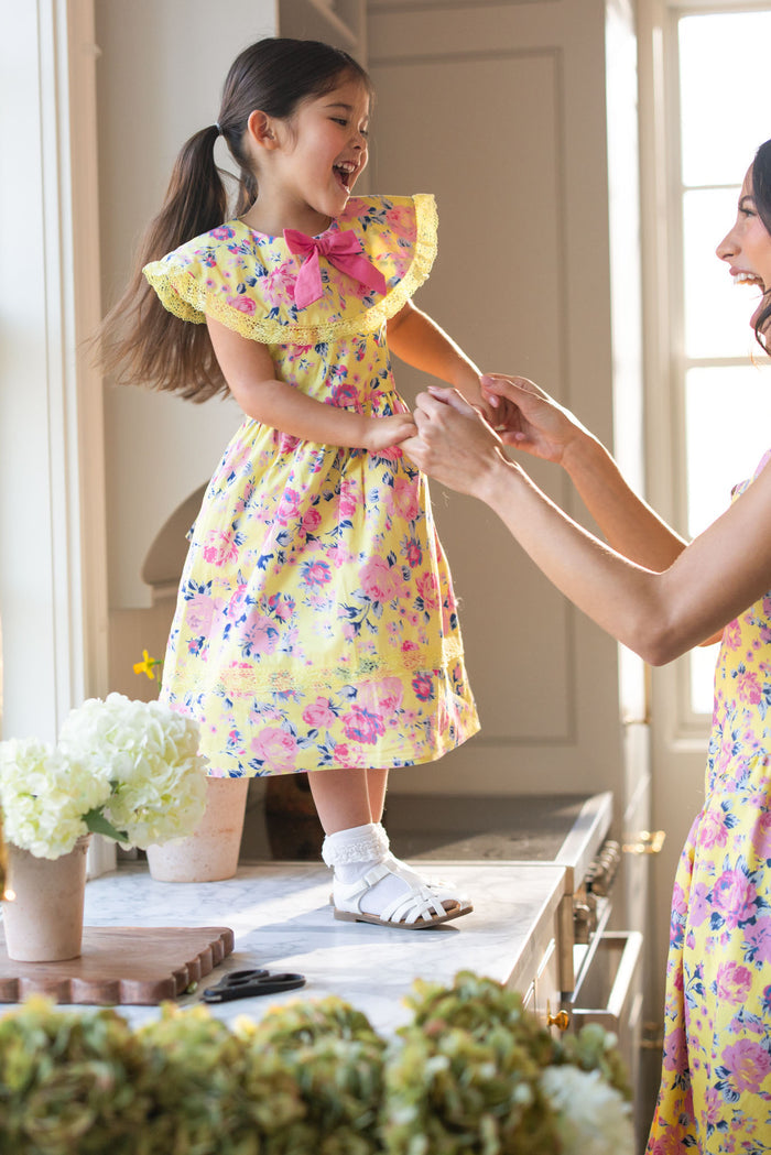 A young girl wears the Ivy City Co Mini Aster Dress in As You Dream Floral, smiling on a kitchen counter as she holds hands with an adult in a matching dress. Sunlight and white flowers brighten the scene.