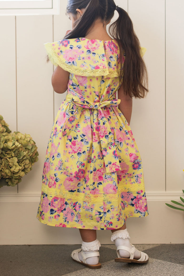 A young girl with long brown hair in pigtails faces away, wearing the Ivy City Co Mini Aster Dress in As You Dream Floral, paired with white socks and sandals. She stands indoors by a wall with plants on the floor.