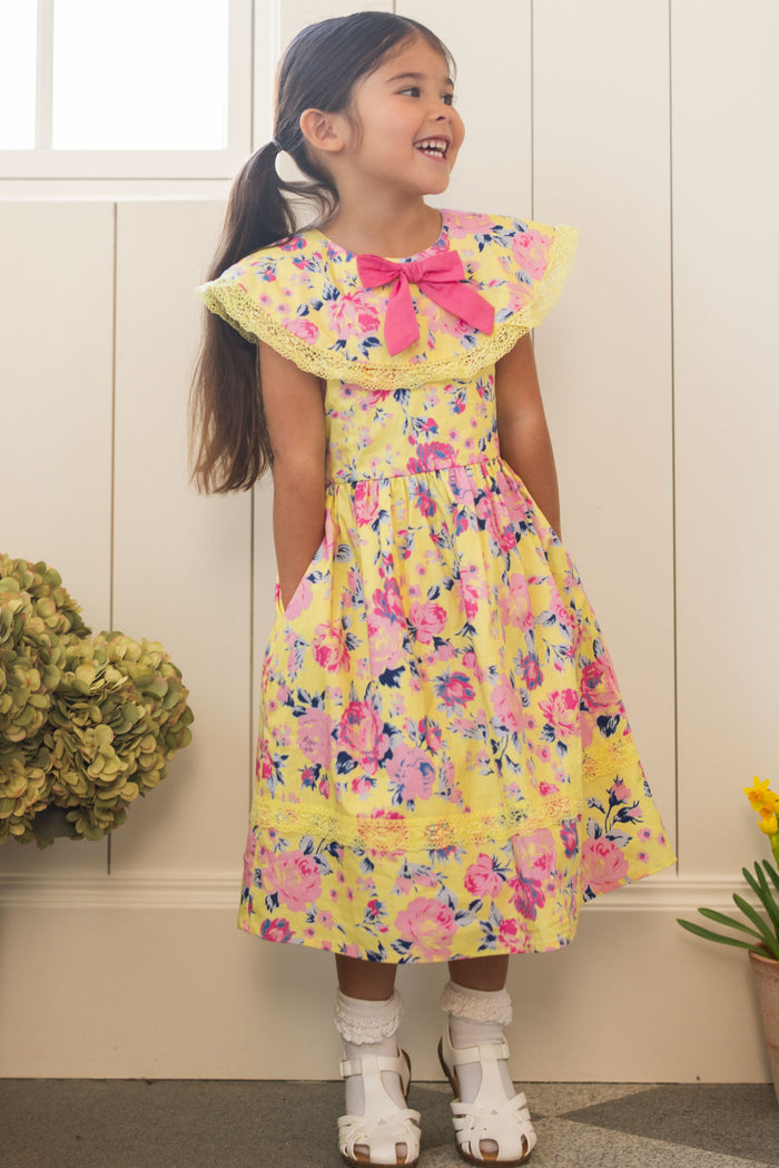 A young girl with long dark hair smiles indoors in the Ivy City Co Mini Aster Dress in As You Dream Floral, featuring a lace-trimmed collar and pink bow at the neck, paired with white sandals. Flowers and a window are behind her.