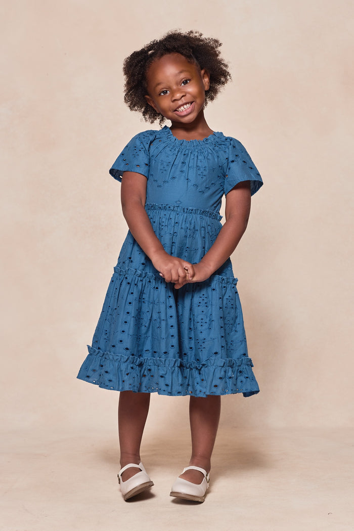 A young girl with curly hair smiles, posing with clasped hands in front of a beige background. She wears the Ivy City Co Mini Etta Dress in Cobalt—a blue, textured cotton dress with short sleeves—paired with white shoes.