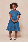 A young girl with curly hair smiles, posing with clasped hands in front of a beige background. She wears the Ivy City Co Mini Etta Dress in Cobalt—a blue, textured cotton dress with short sleeves—paired with white shoes.