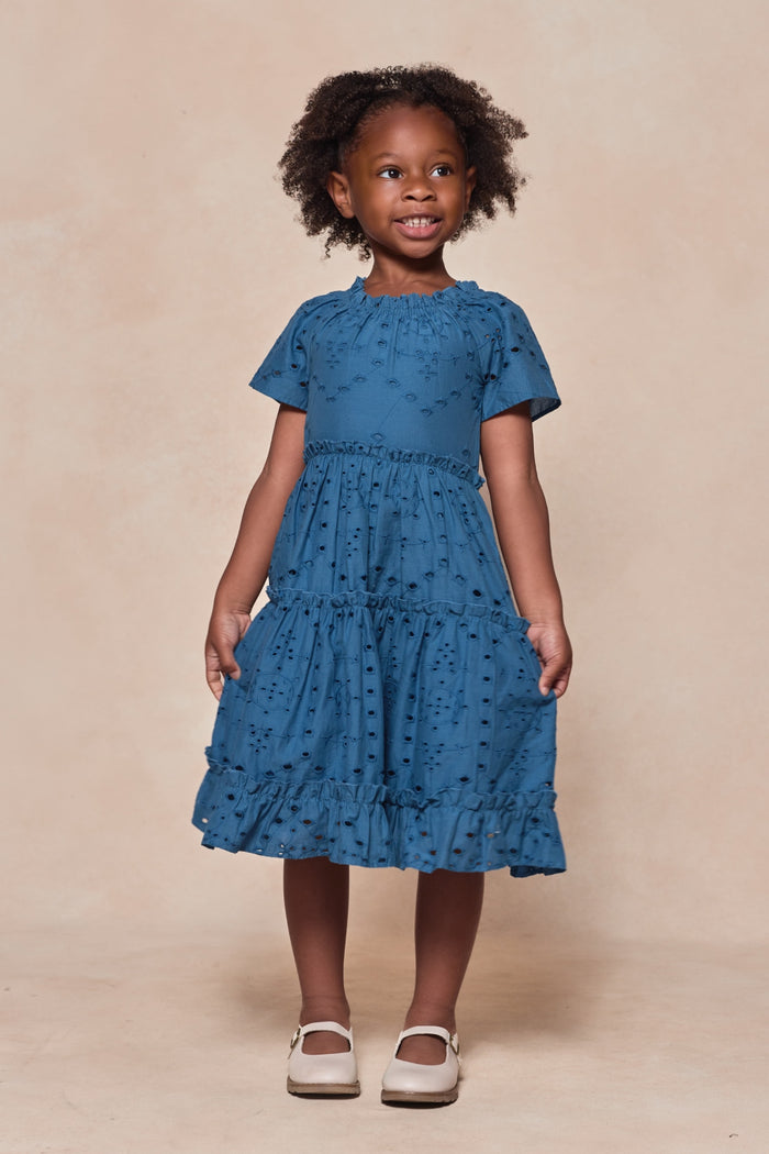 A young girl with curly hair smiles in the Ivy City Co Mini Etta Dress in Cobalt and white shoes, standing against a beige background.