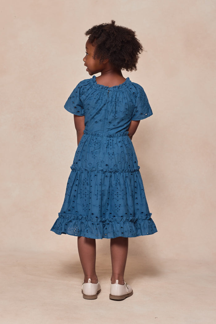 A young girl faces away, wearing Ivy City Co's Mini Etta Dress in Cobalt—a short-sleeved, knee-length cotton dress with ruffled layers—paired with white shoes, set against a light beige background.