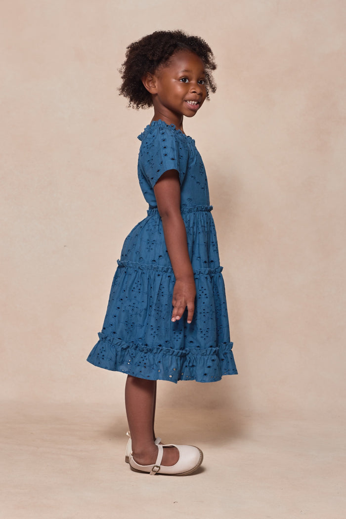 A young girl with curly hair smiles sideways, wearing the Mini Etta Dress in Cobalt by Ivy City Co. The cotton dress features ruffles and eyelet details. She pairs it with white shoes, posing against a beige background.