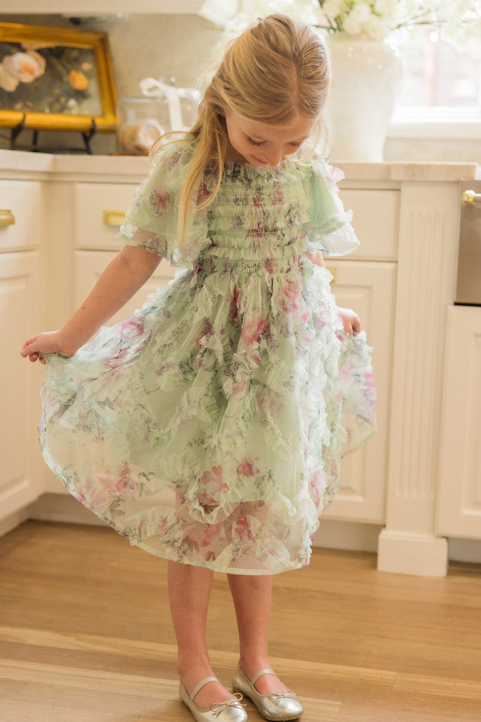 A young girl with long blonde hair holds out the sides of her Ivy City Co Mini Florentina Dress in Pistachio Aquarelle Fleur, featuring a smocked bodice, while standing in a bright kitchen with white cabinets and wooden floors.