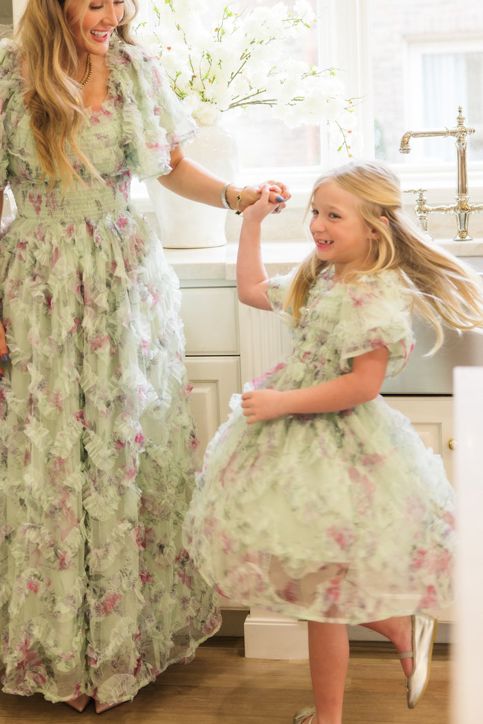 A woman and young girl smile and hold hands while dancing in a bright kitchen, both wearing matching Ivy City Co Florentina Dresses in Pistachio Aquarelle Fleur. White flowers are visible by the window.
