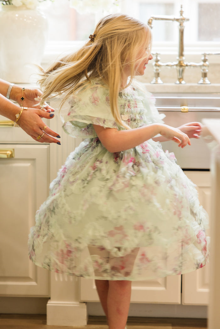 A young girl with long blonde hair twirls and smiles in a bright kitchen, wearing the Ivy City Co Mini Florentina Dress in Pistachio Aquarelle Fleur, as an adult’s hands reach toward her.