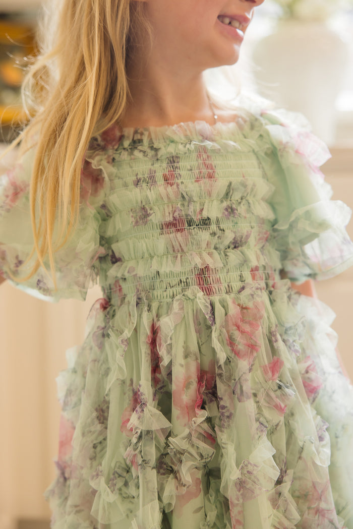 A young girl smiles, wearing the Ivy City Co Mini Florentina Dress in Pistachio Aquarelle Fleur, featuring a smocked bodice and ruffled details. Her loose blonde hair and a bright background create a cheerful, sunlit indoor scene.