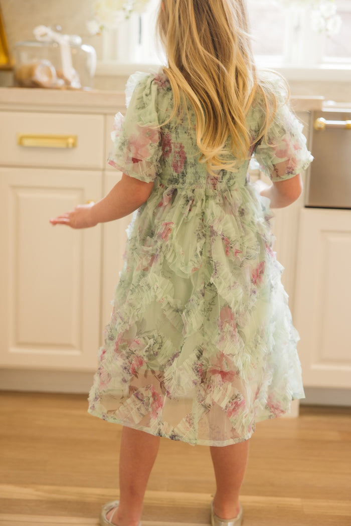 A young girl with long blond hair stands barefoot in a bright kitchen, wearing the Ivy City Co Mini Florentina Dress in Pistachio Aquarelle Fleur, facing away from the camera.