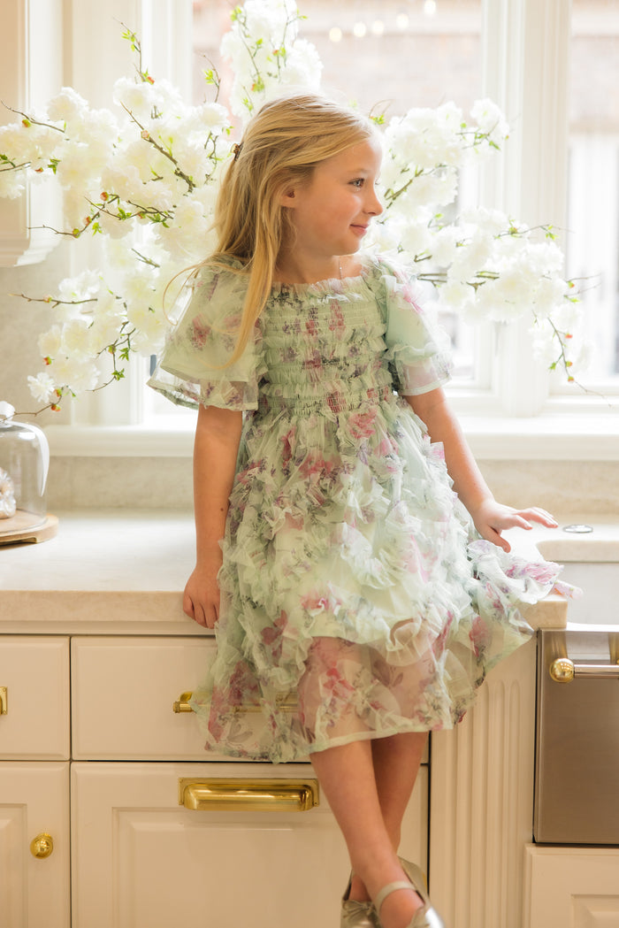A young girl with long blonde hair, smiling as she sits on a kitchen counter, wears the Ivy City Co Mini Florentina Dress in Pistachio Aquarelle Fleur. Sunlight and white flowers brighten the space behind her.