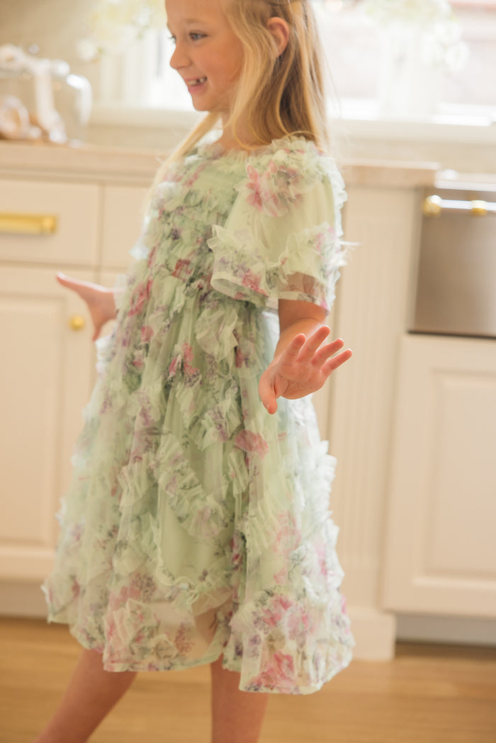 A young girl in the Ivy City Co Mini Florentina Dress in Pistachio Aquarelle Fleur stands smiling in a sunlit kitchen, holding her hands out playfully as light streams through the window behind her.