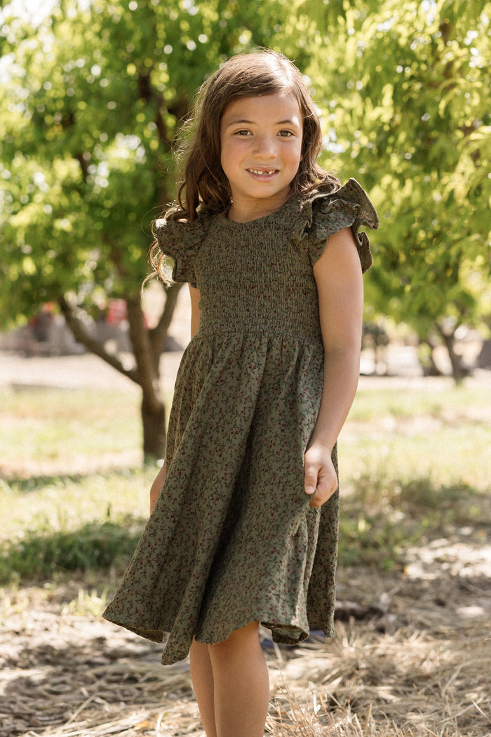 A young girl with long brown hair smiles while standing outside among green trees on a sunny day, wearing the Ivy City Co Mini Hattie Dress in Woodland Floral, featuring ruffled sleeves and a smocked bodice.