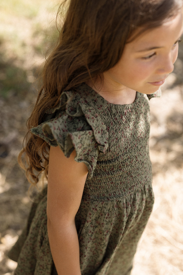 A young girl with long brown hair stands outdoors on a sunny day, looking to the side, wearing the Ivy City Co Mini Hattie Dress in Woodland Floral.
