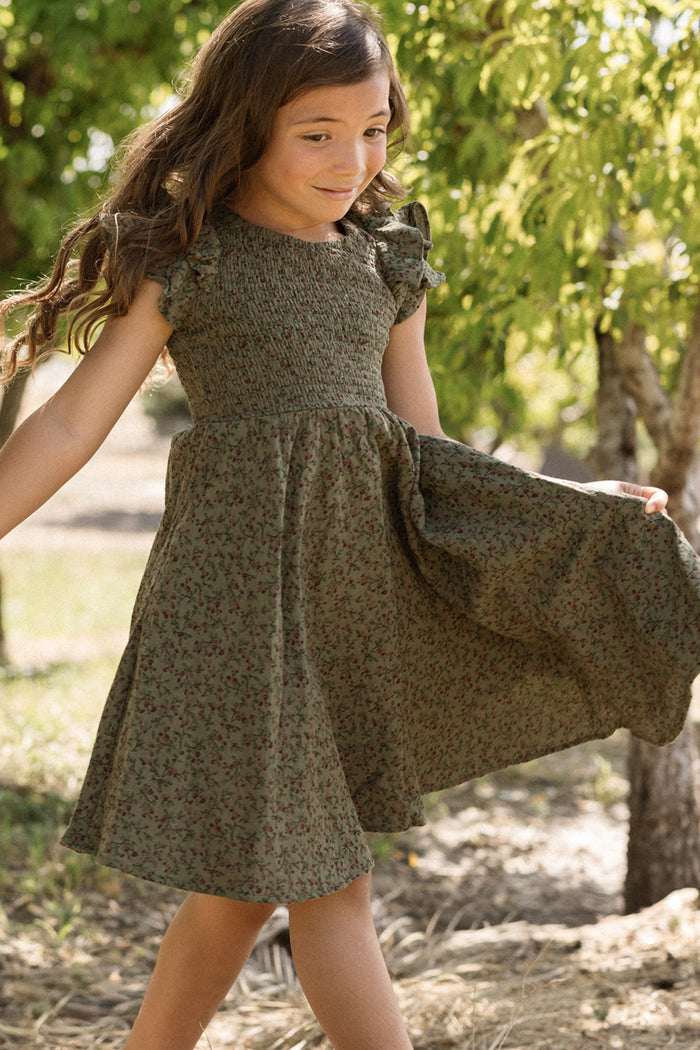 A smiling young girl with long brown hair, wearing the Ivy City Co Mini Hattie Dress in Woodland Floral with a smocked bodice, walks outdoors among trees on a sunny day.