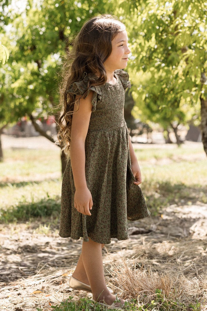 A young girl with long brown hair stands outdoors in the Mini Hattie Dress in Woodland Floral by Ivy City Co, its smocked bodice lending charming detail to the cotton dress. She smiles and looks off to the side, surrounded by leafy trees on a sunny day.