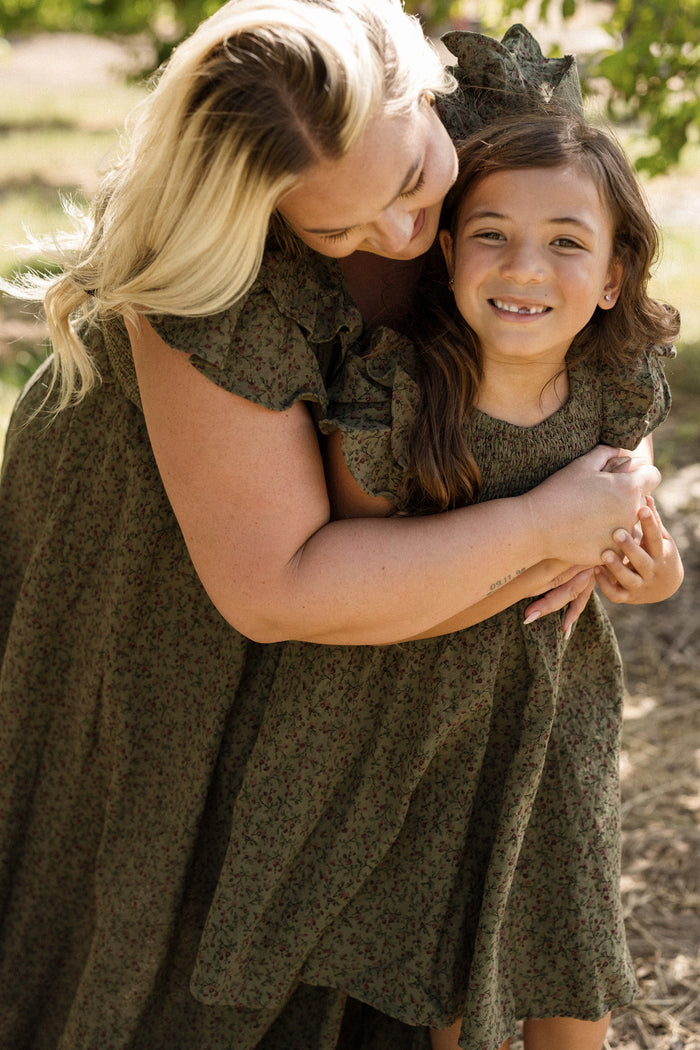 A woman hugs a smiling young girl outdoors, both wearing the Ivy City Co Mini Hattie Dress in Woodland Floral with a smocked bodice. Sunlight filters through the trees in the background.