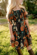 A young girl with long blonde hair wears the Ivy City Co Mini Madeline Dress in Imogen Moody Floral, a charming fall floral dress paired with brown boots, standing on green grass in sunlight, with trees and a fence softly blurred in the background.