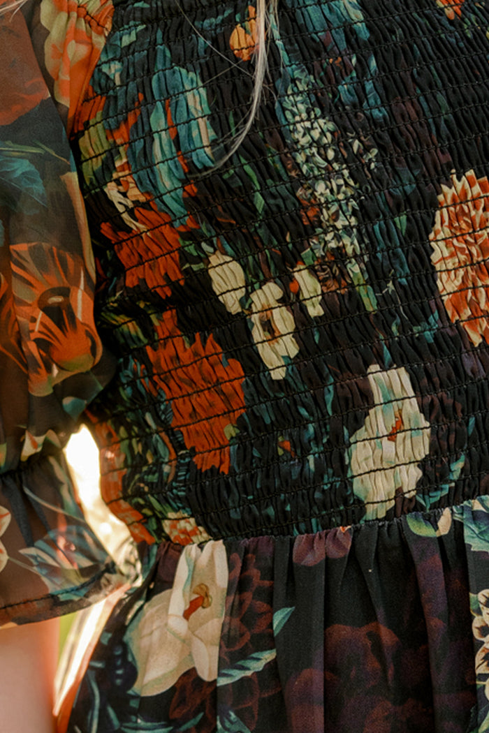 Close-up of a person wearing the Ivy City Co Mini Madeline Dress in Imogen Moody Floral, a black chiffon dress adorned with orange, green, and white florals. The dress features smocked fabric on the bodice and sheer puff sleeves—making it an ideal stylish choice for fall.