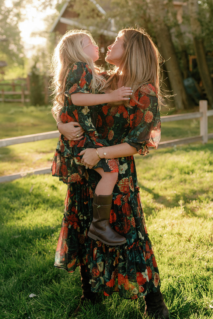 A woman and a young girl, both dressed in matching Ivy City Co Mini Madeline Dresses in Imogen Moody Floral, stand together in a sunlit yard. The woman holds the girl as they share a smile, with trees and a wooden fence visible in the background.