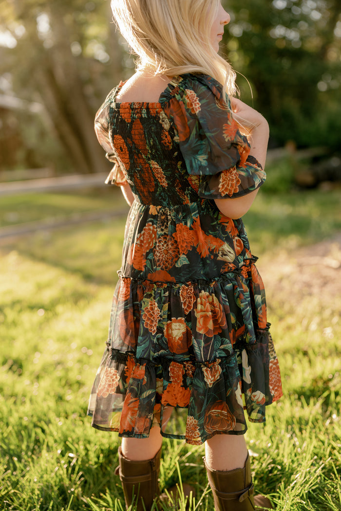 A child with long blonde hair stands on grass wearing the Ivy City Co Mini Madeline Dress in Imogen Moody Floral, paired with brown boots. The dark floral dress features orange flowers and ruffles, while sunlight casts a warm, golden glow in the background, perfectly showcasing this charming fall piece.