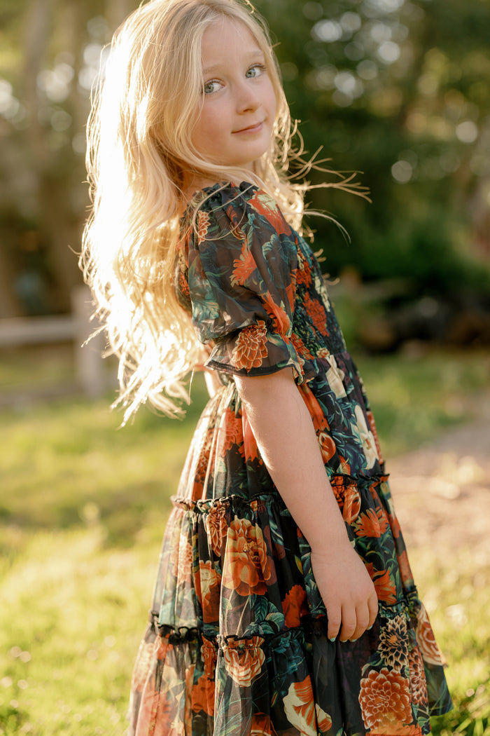 A young girl with long blonde hair stands outdoors in sunlight, wearing the Mini Madeline Dress in Imogen Moody Floral by Ivy City Co— a dark chiffon dress adorned with orange and green floral patterns. She looks over her shoulder with a gentle expression, set against a backdrop of blurred greenery.