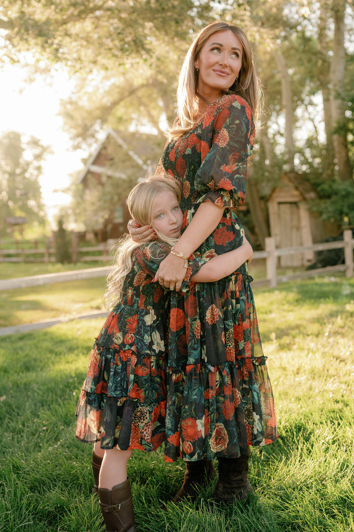A woman and a young girl, both dressed in matching chiffon outfits, stand together in a sunlit grassy yard. The girl hugs the woman's waist while she gazes ahead, with trees and a wooden fence in the background—a perfect scene to highlight the charming Ivy City Co Mini Madeline Dress in Imogen Moody Floral for fall.