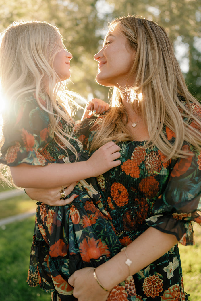 A woman and a young girl, both wearing the Ivy City Co Mini Madeline Dress in Imogen Moody Floral, smile at each other in warm sunlight as they stand on green grass with trees in the background.