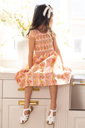 A young girl with long dark hair, wearing a white hair bow and Ivy City Co's Mini Marlow Cap Sleeve Dress in Peach Handpainted Flora Stripe, sits on a sunlit kitchen counter near a vase of white flowers, looking to the side in white sandals.