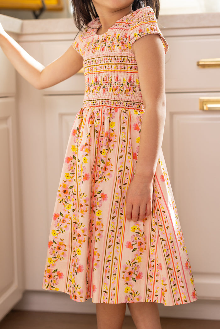 A young girl stands in a kitchen wearing the Ivy City Co Mini Marlow Cap Sleeve Dress in Peach Handpainted Flora Stripe. The short-sleeved dress features floral details and vertical striped flower patterns. Her face is not visible.