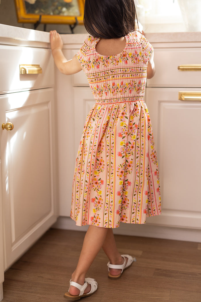 A young girl in the Ivy City Co Mini Marlow Cap Sleeve Dress in Peach Handpainted Flora Stripe and white sandals stands in a bright kitchen, her back to the camera as sunlight streams through a window.