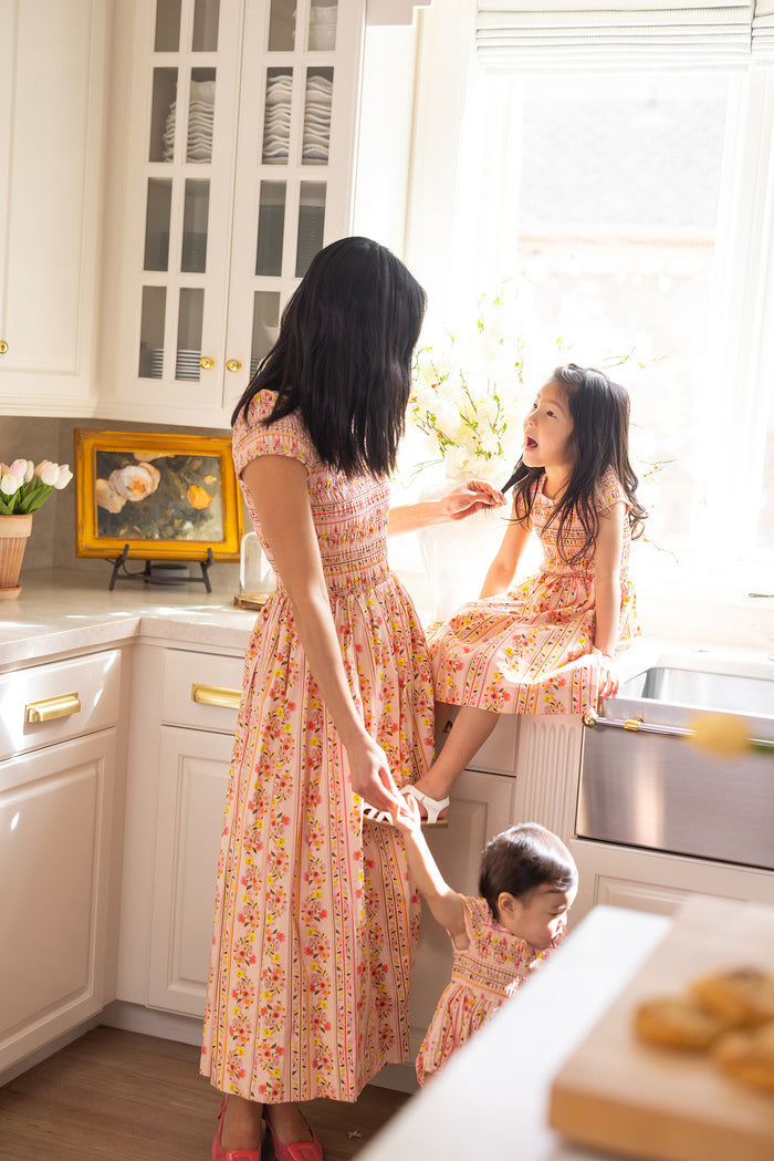 A woman and two young girls wear matching Ivy City Co Marlow Cap Sleeve Dresses in Peach Handpainted Flora Stripe as they enjoy a bright kitchen moment, with one girl at the sink and the other holding hands for a sweet spring scene.
