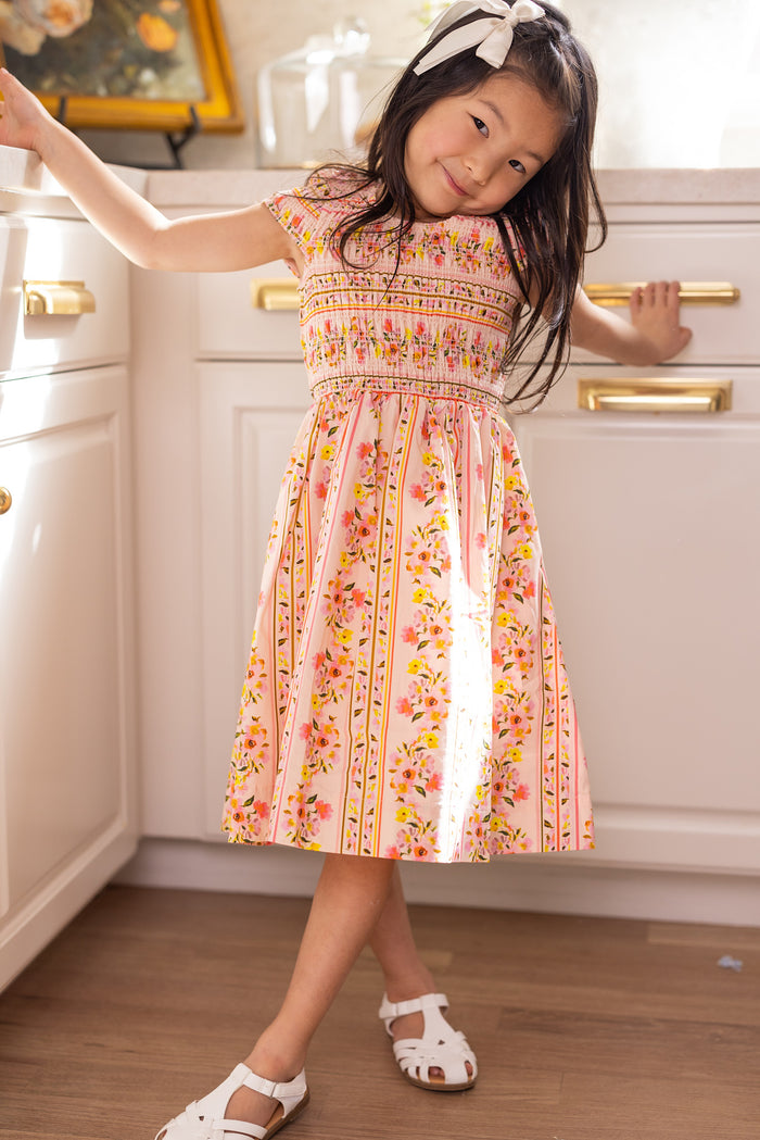 A young girl smiles in a sunlit kitchen, wearing the Ivy City Co Mini Marlow Cap Sleeve Dress in Peach Handpainted Flora Stripe, paired with white sandals and a white bow headband.