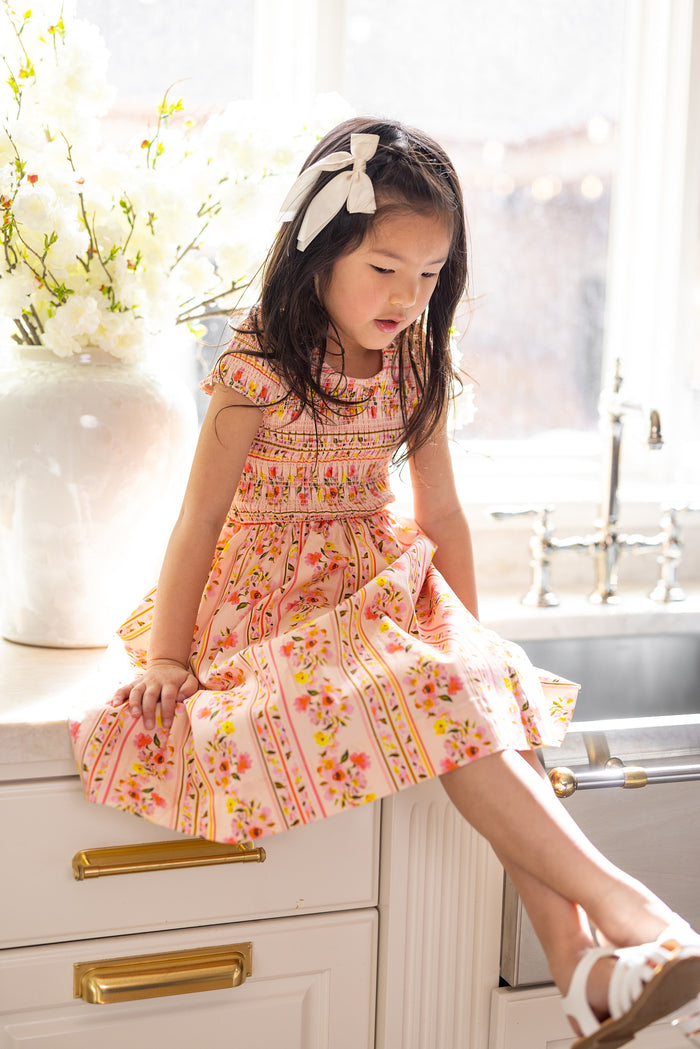 A young girl wears the Ivy City Co Mini Marlow Cap Sleeve Dress in Peach Handpainted Flora Stripe, sitting on a kitchen counter by the sink with sunlight streaming in and a vase of white flowers glowing softly in the background.