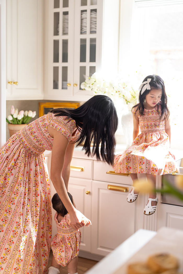 A woman in the Ivy City Co Mini Marlow Cap Sleeve Dress in Peach Handpainted Flora Stripe assists a toddler while a young girl, also in a matching dress, sits on the kitchen counter with white cabinets and fresh flowers.