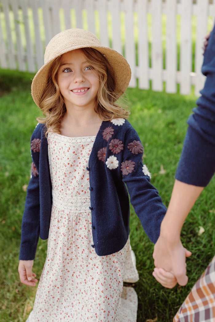A smiling girl wearing Ivy City Co’s Mini Pauline Sweater in Midnight Floral over her white floral dress holds hands outdoors near a white picket fence.
