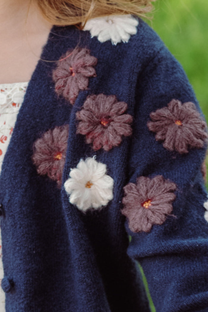 An Ivy City Co Mini Pauline Sweater in Midnight Floral, featuring large knitted flower appliqués in white and purple, styled over a patterned white dress.