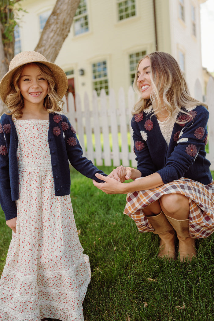 A woman and young girl smile outdoors by a white picket fence. Both wear navy cardigans with floral patterns—one resembling an embroidered sweater, and the other, Ivy City Co’s charming Mini Pauline Sweater in Midnight Floral. The girl also wears a straw hat. They hold hands, sitting and standing on green grass.