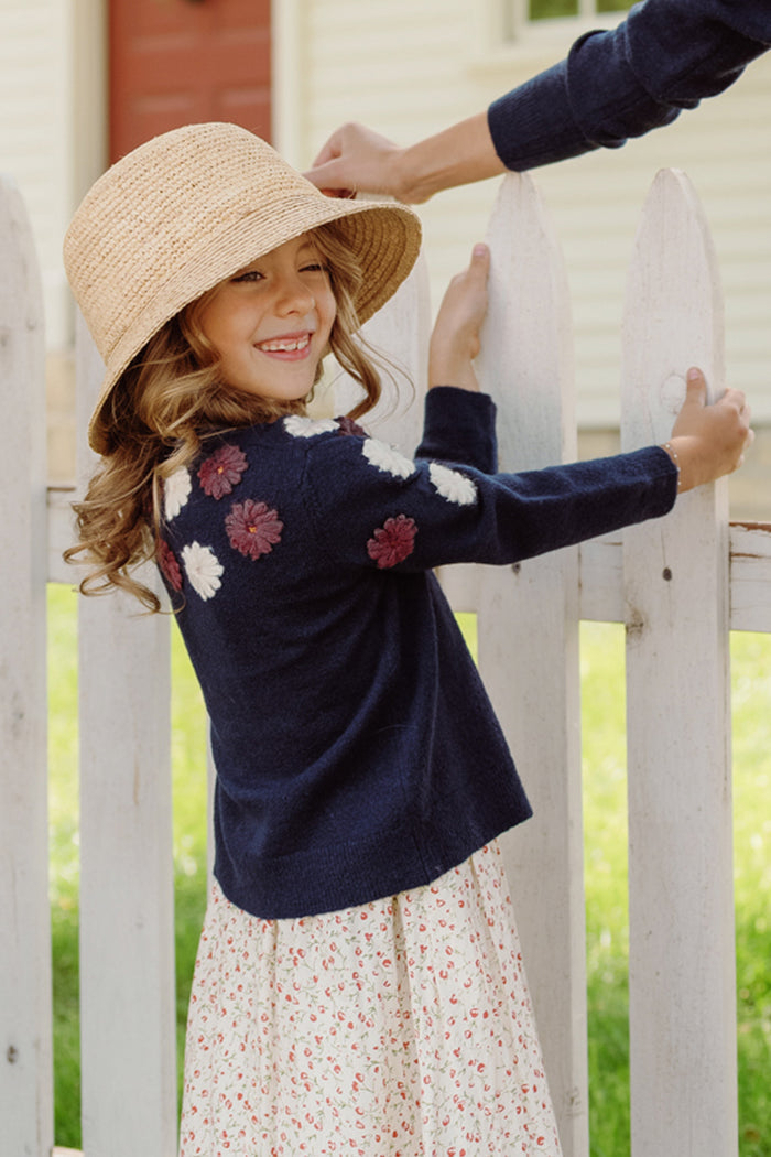 A smiling young girl in a straw hat and the Mini Pauline Sweater in Midnight Floral by Ivy City Co stands by a white picket fence, holding onto a fence post, with another person’s arm visible in the background.