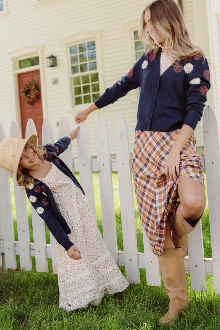 A woman and a young girl stand by a white picket fence, both dressed in floral cardigans and dresses. The girl smiles brightly in her Ivy City Co Mini Pauline Sweater in Midnight Floral, accessorized with a straw hat, while the woman completes her look with boots. A cream house with windows serves as the backdrop.