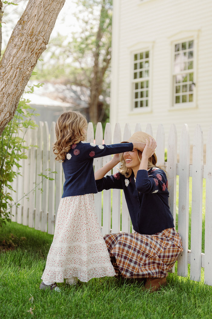 A young girl playfully places a straw hat on a smiling adult’s head. Both are dressed in flowered sweaters—one wearing the Ivy City Co Mini Pauline Sweater in Midnight Floral, and the other in an embroidered sweater—paired with patterned dresses. They stand by a white picket fence, surrounded by grass, trees, and a house in the background.