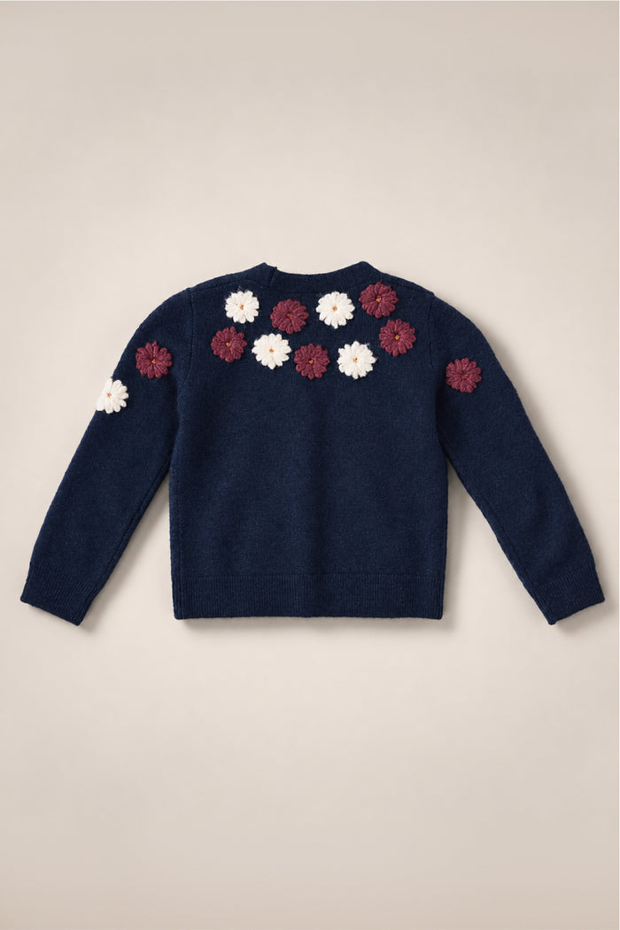 The Mini Pauline Sweater in Midnight Floral by Ivy City Co is a navy blue v-neck sweater with long sleeves, adorned with white and maroon floral appliqués on the upper back and shoulders. Displayed against a light beige background, this embroidered piece is part of the Mini Pauline Sweater collection.