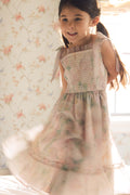 A young girl with long dark hair smiles as she twirls in the Ivy City Co Mini Rhiannon Dress in Painted Bloom, featuring a smocked bodice. She stands in a bright, flower-wallpapered room, ready for any special occasion.