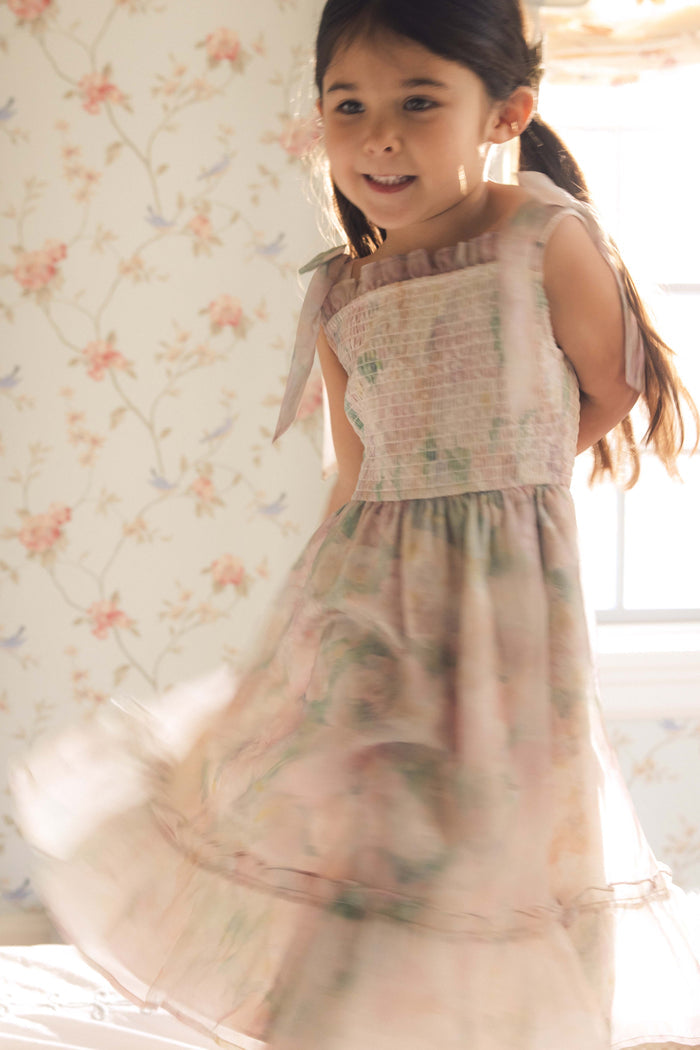 A young girl with long dark hair smiles as she twirls in the Ivy City Co Mini Rhiannon Dress in Painted Bloom, featuring a smocked bodice. She stands in a bright, flower-wallpapered room, ready for any special occasion.