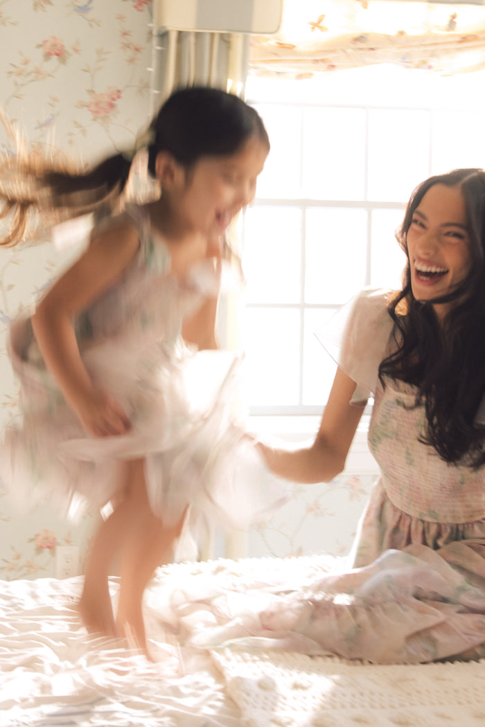 A woman and a young girl laugh together as the girl jumps on a bed in her Ivy City Co Mini Rhiannon Dress in Painted Bloom, perfect for special occasions, in a sunlit room with floral wallpaper and a window behind.