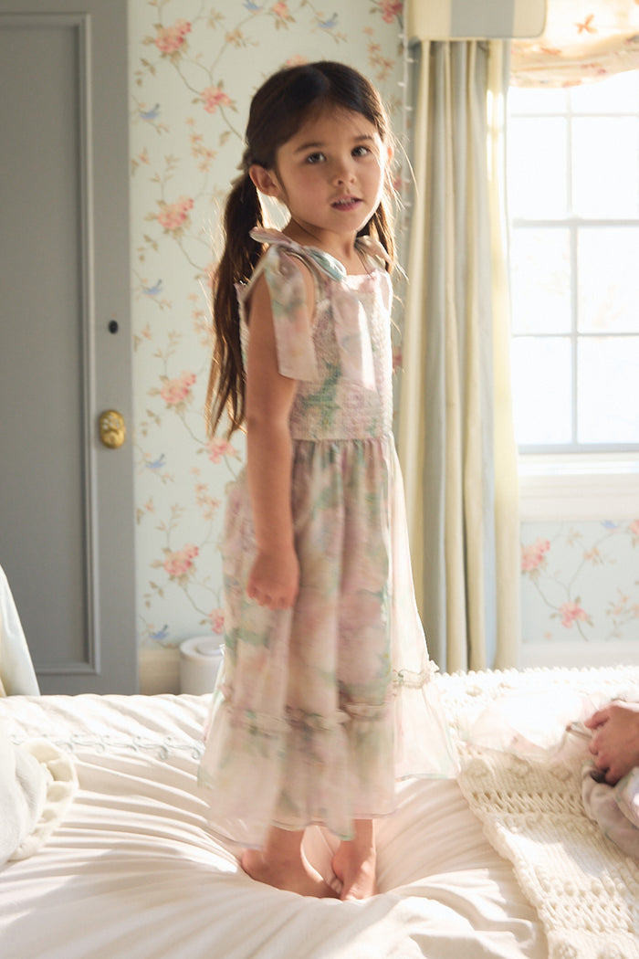 A young girl with long brown hair in pigtails stands barefoot on a bed, wearing the Mini Rhiannon Dress in Painted Bloom by Ivy City Co. The softly lit room has floral wallpaper and a window with curtains.