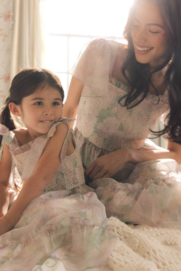 A woman and a young girl sit together on a bed in soft light. The girl, content, wears the Ivy City Co Mini Rhiannon Dress in Painted Bloom, which features watercolor floral organza and a smocked bodice.