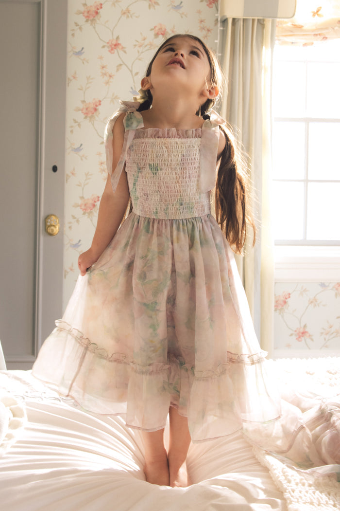 A young girl in the Ivy City Co Mini Rhiannon Dress in Painted Bloom, featuring a smocked bodice, stands on a bed and smiles as sunlight spills through the window into a room with floral wallpaper and light curtains.